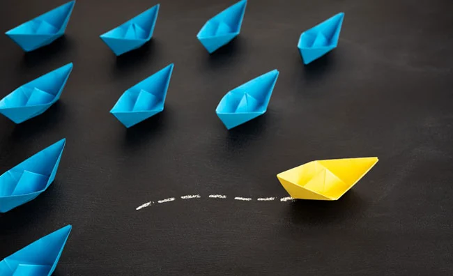 A single yellow paper boat leading a group of six blue paper boats on a dark surface, with a dashed white line trailing behind the yellow boat.