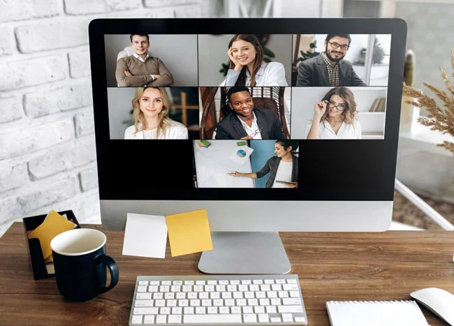 A computer monitor on a wooden desk showing a group video call with seven diverse professionals in a grid view, next to a coffee mug and sticky notes.