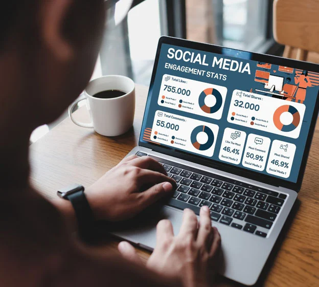 An over-the-shoulder view of a person using a laptop that displays a "Social Media Engagement Stats" dashboard featuring colorful donut charts, numerical data, and growth metrics.