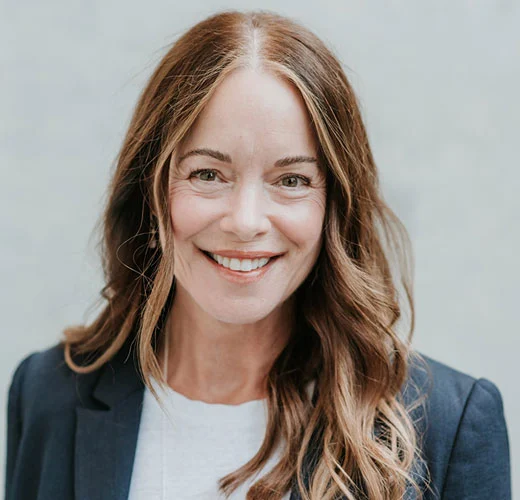 Smiling professional woman with wavy brown hair wearing a navy blue blazer and white t-shirt.