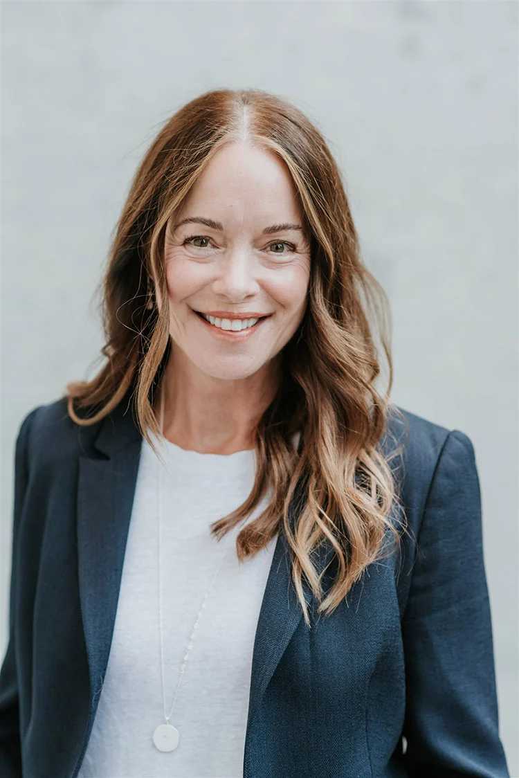 Smiling professional woman with wavy brown hair wearing a navy blue blazer and white t-shirt.