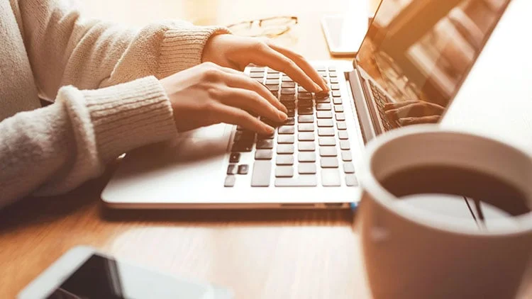 Close-up of hands in a white sweater typing on a laptop next to a cup of coffee in warm sunlight.