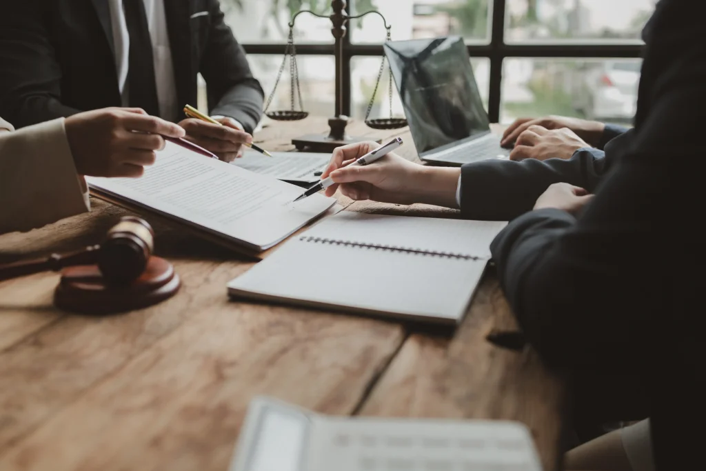A group of legal professionals in suits sitting around a rustic wooden table, reviewing and signing a document with a gavel and scales of justice in the background.