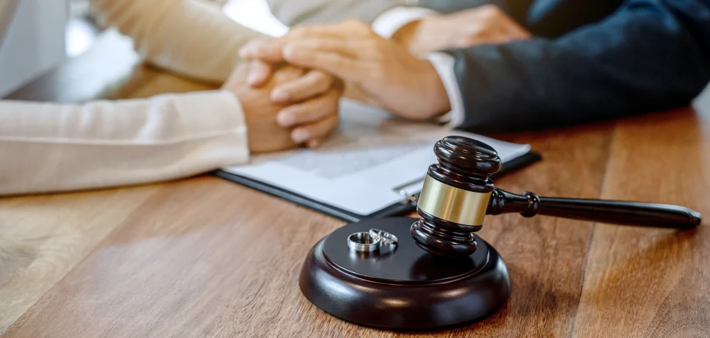 A close-up shot of two people holding hands over a legal document on a wooden desk, with a wooden gavel and two silver wedding rings in the foreground.